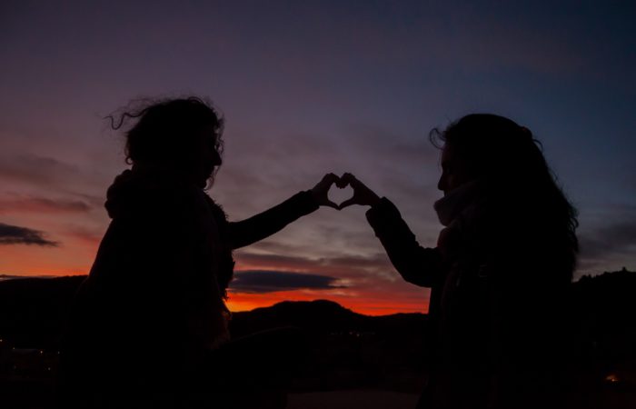 Two women joining their hands together to form a heart, with the sunset behind them