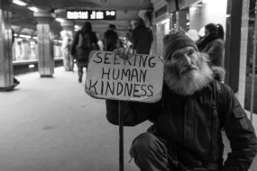 A black and white picture of a man in a subway holding up a sign saying "Seeking Human Kindness"