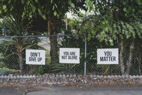 Signs behind a chain-link fence that say "DON'T GIVE UP," "YOU ARE NOT ALONE," "YOU MATTER"