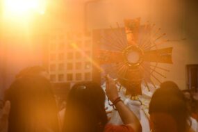 A priest holding up a monstrance to three worshippers, with the sun reflecting brightly off the monstrance