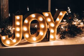 A light-up sign displaying the word "JOY" in the middle of Christmas decorations of pinecones and holly