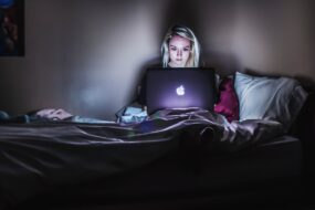 A young woman sitting in a darkened room late at night staring intently at a MacBook which gives off most of the light in the room