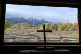 A photo by Mark Roberts of a cross on a windowsill with the Grand Tetons seen through the window