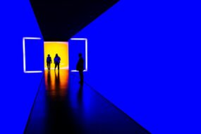 People standing in a doorway of a room; the room is lit in dark blue, but the space beyond the door looks like a beautiful sunrise
