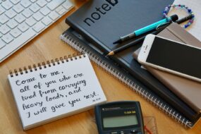 A desk with notebooks, phone, and calculator; on one notebook is written "Come to me, all of you who are tired from carrying heavy loads, and I will give you rest" (Matthew 11:28)
