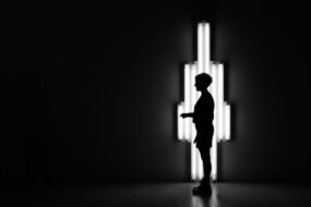 A black and white photo of a person standing alone in a room with harsh lights behind them
